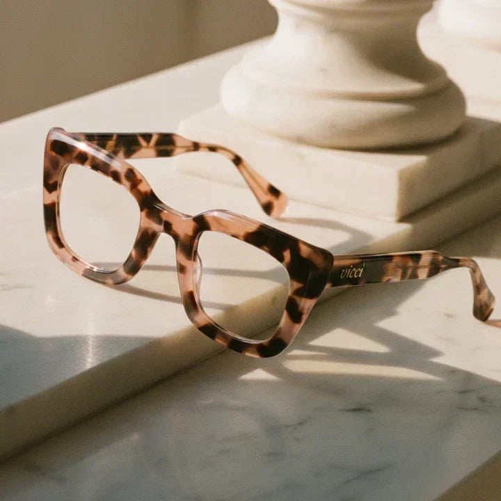 Vicci Eyewear’s Sofia cat-eye frames in pink tortoise, resting on a sunlit marble surface with sculptural background detail
